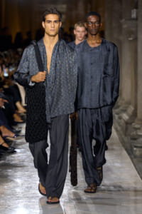 Two male models walk a fashion runway in dark blue-gray outfits; one carries a large black knitted bag over his shoulder.