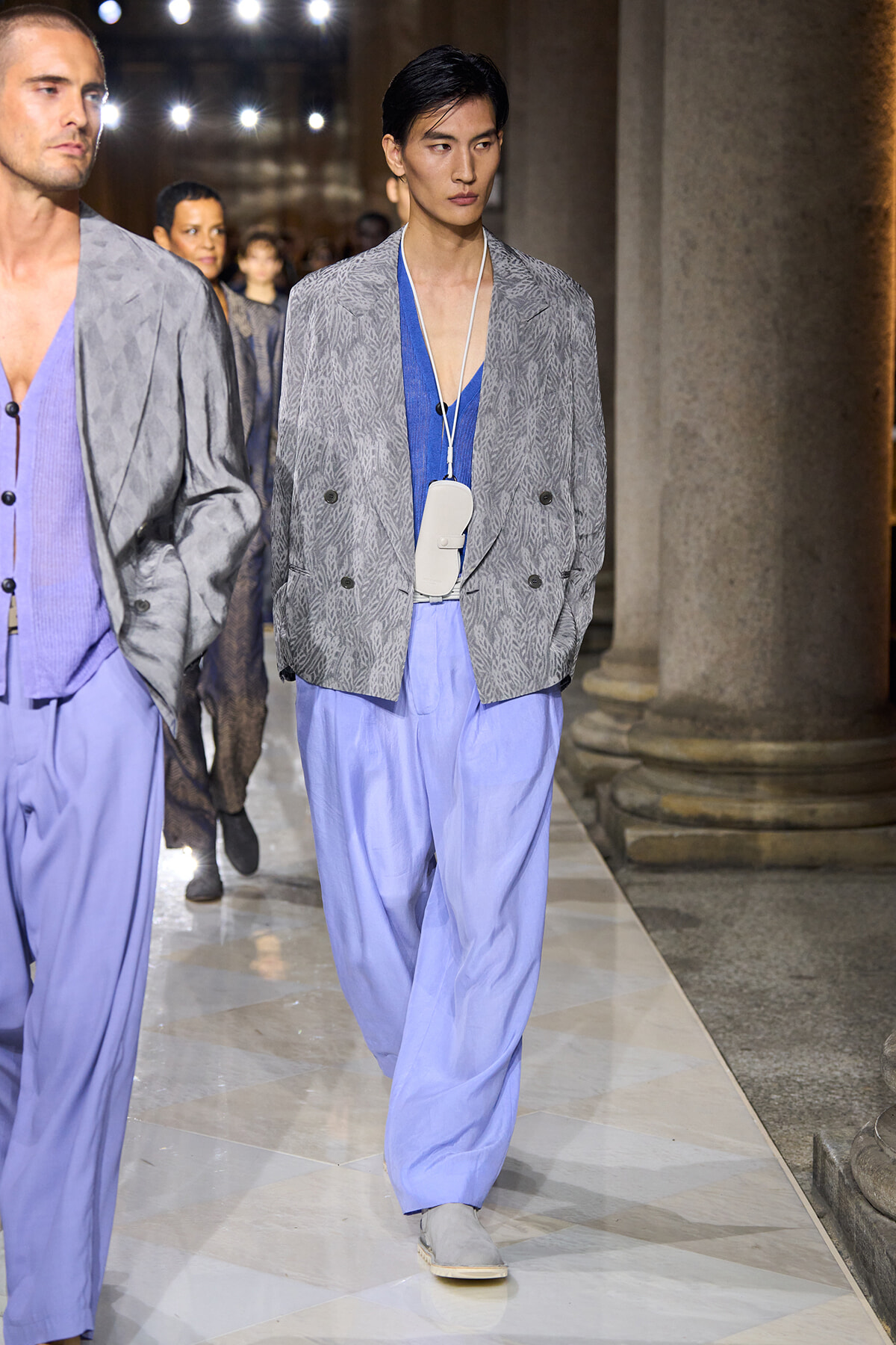 Male model walks a runway in a gray patterned blazer over a blue top with lavender wide-leg pants and a white pendant accessory.