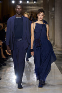 Two models walk a fashion show runway in navy blue outfits: a tall man in a relaxed suit and a woman in a flowing dress with a small crossbody bag under bright lights start the line.