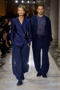 Two models walk a fashion show in coordinated navy outfits; woman wears an oversized navy blazer with wide-pleated pants and blue footwear, man wears a navy shirt with cardigan and matching pants.