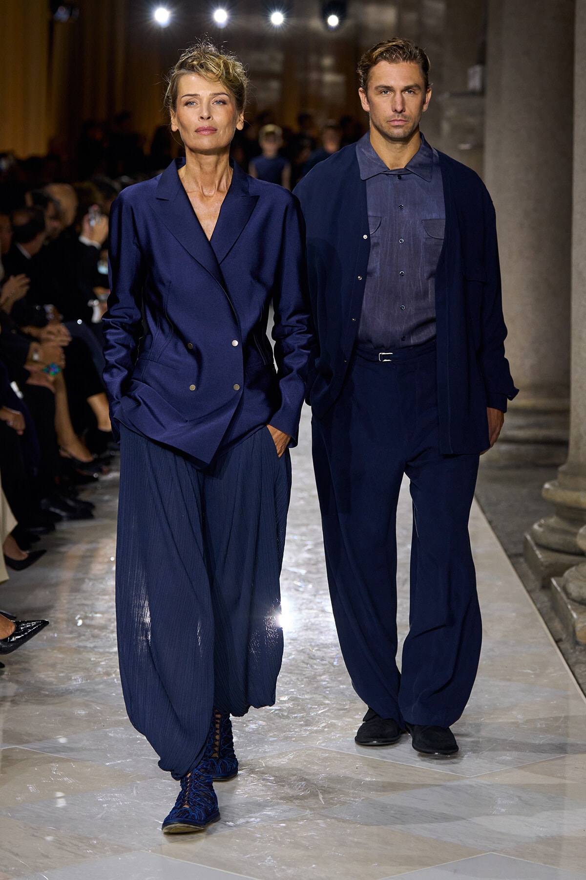 Two models walk a fashion show in coordinated navy outfits; woman wears an oversized navy blazer with wide-pleated pants and blue footwear, man wears a navy shirt with cardigan and matching pants.