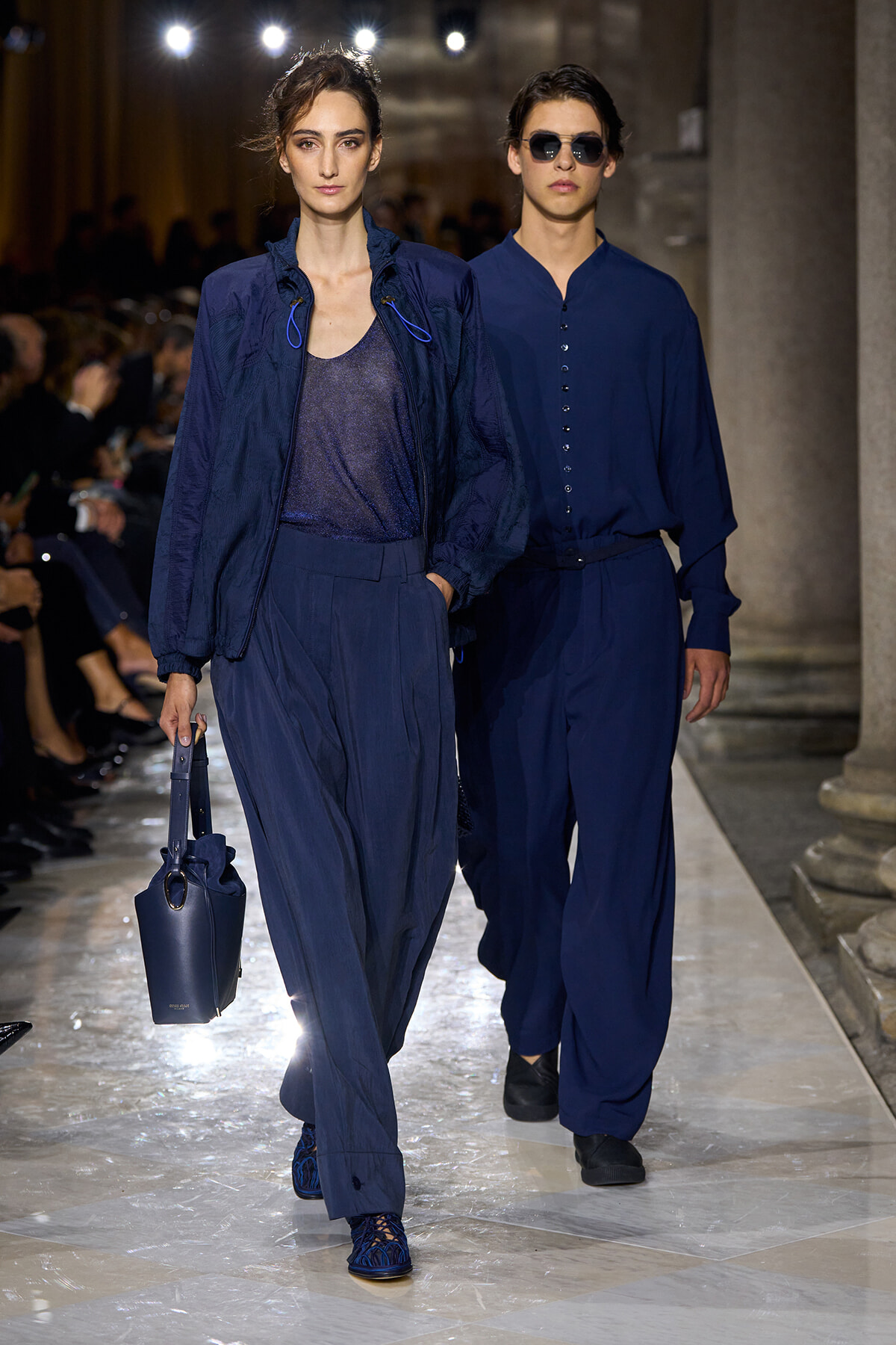 Two models walk a runway in navy blue outfits; left model wears a sheer top with a bomber jacket and wide-leg pants, carrying a navy bag with a ring handle on the side service.