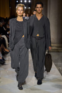 Two models walk a runway in gray outfits: a woman in a draped blouse and blazer, a man in a layered gray vest and jacket, with a brown woven bag in hand (fashion show).