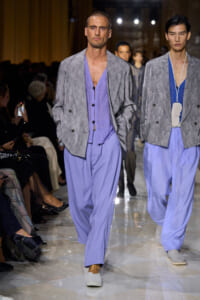 Male models walk a fashion show runway in lavender outfits: loose lavender trousers and vests under gray patterned jackets, with audience seated along the sides.
