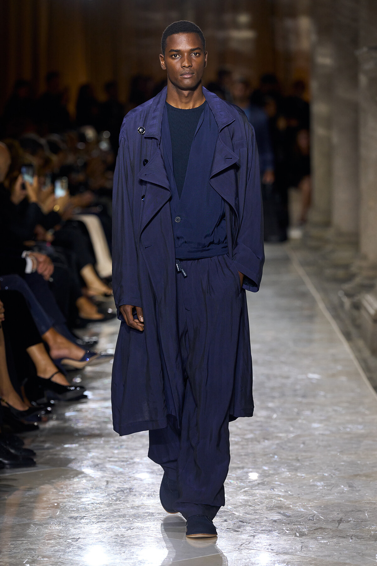 Male model walks a runway wearing a loose navy trench coat over a dark outfit.