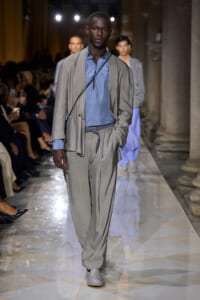 Male model walks a fashion runway in a light gray suit with a blue shirt and a crossbody strap.