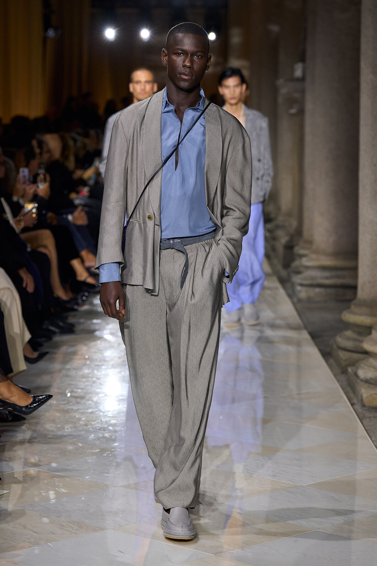 Male model walks a fashion runway in a light gray suit with a blue shirt and a crossbody strap.