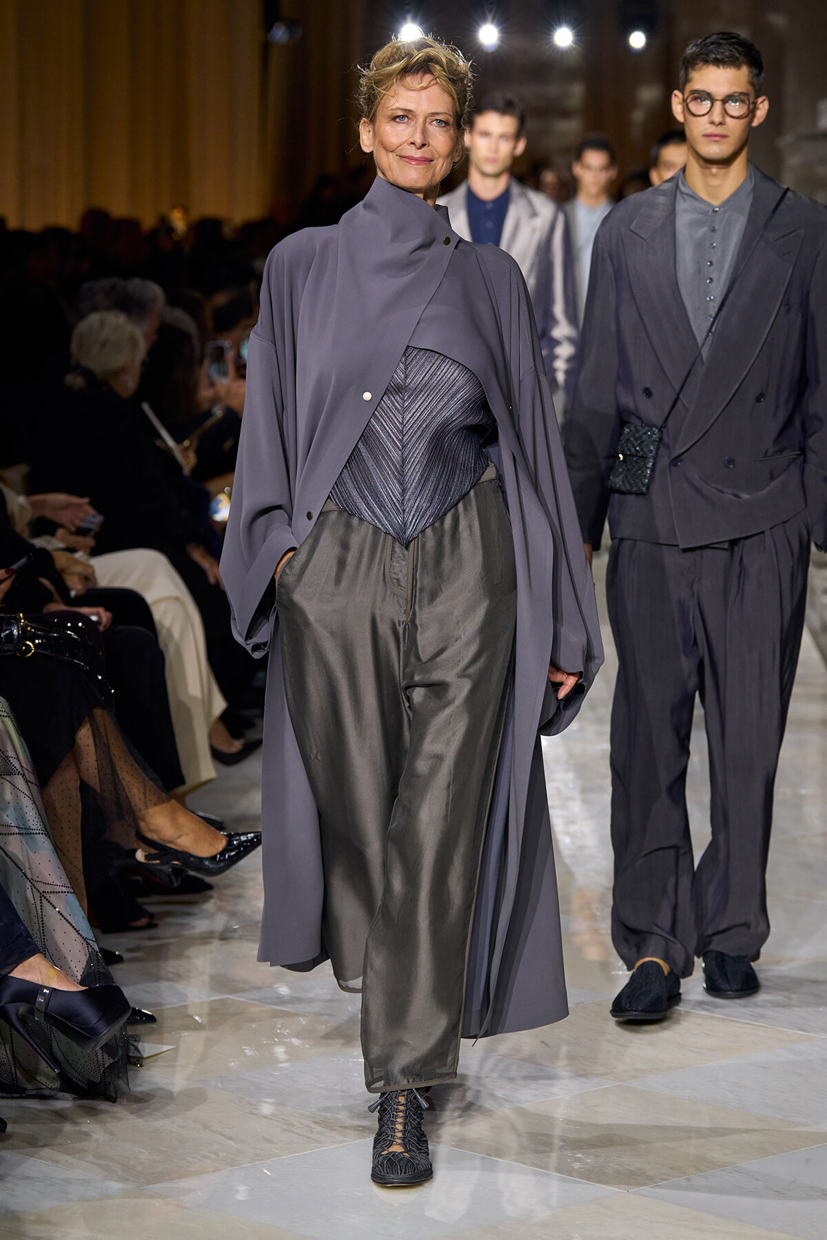Fashion model walking a runway in a gray layered ensemble: a cape over a metallic bodice and wide gray pants, with audience in the background.