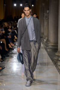 Male fashion model walking a runway in a gray blazer over a light gray shirt, patterned loose pants, and gray shoes, holding a blue hat in one hand.