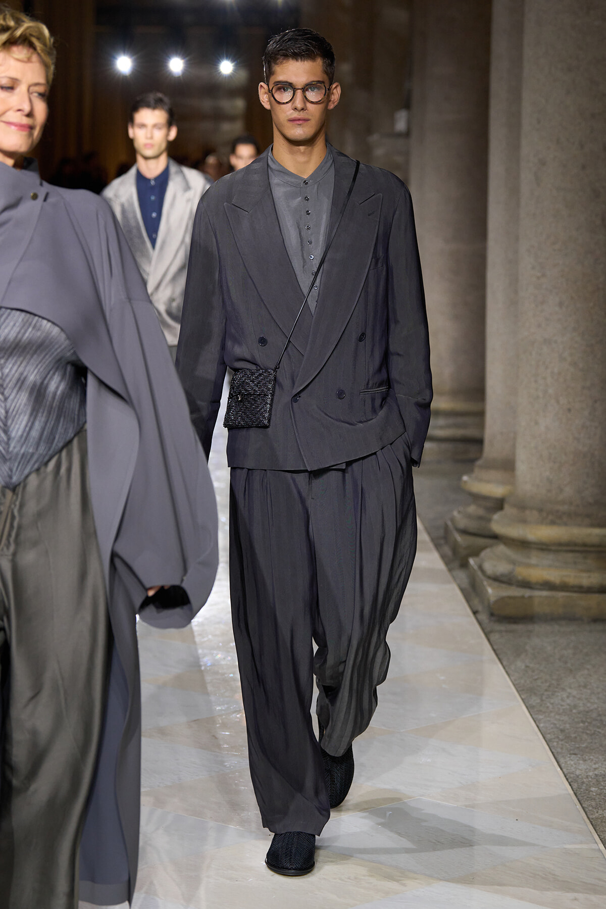Male model on a runway wearing a dark gray oversized suit, double-breasted blazer, wide trousers, round glasses, and a small black crossbody bag.