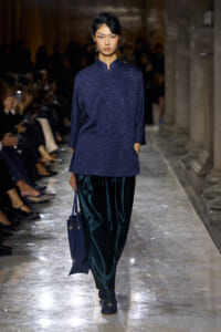 Female model walking a runway in a navy textured top and dark teal velvet pants, carrying a navy bag.