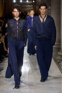 Two models walk a runway in navy-blue outfits: a sheer, lattice-knit jacket over wide-leg pants and a matching bag on the left; a male model in a textured navy cardigan with a tie and wide trousers on the right.