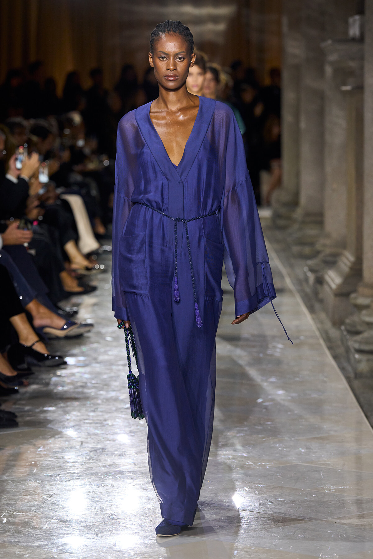 Runway model wears a cobalt blue sheer-sleeve jumpsuit with a wrapped belt and tassel accessories.