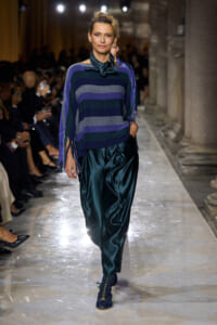 Model on a runway wearing a striped purple and blue poncho with fringe, teal satin pants, and matching lace-up teal shoes