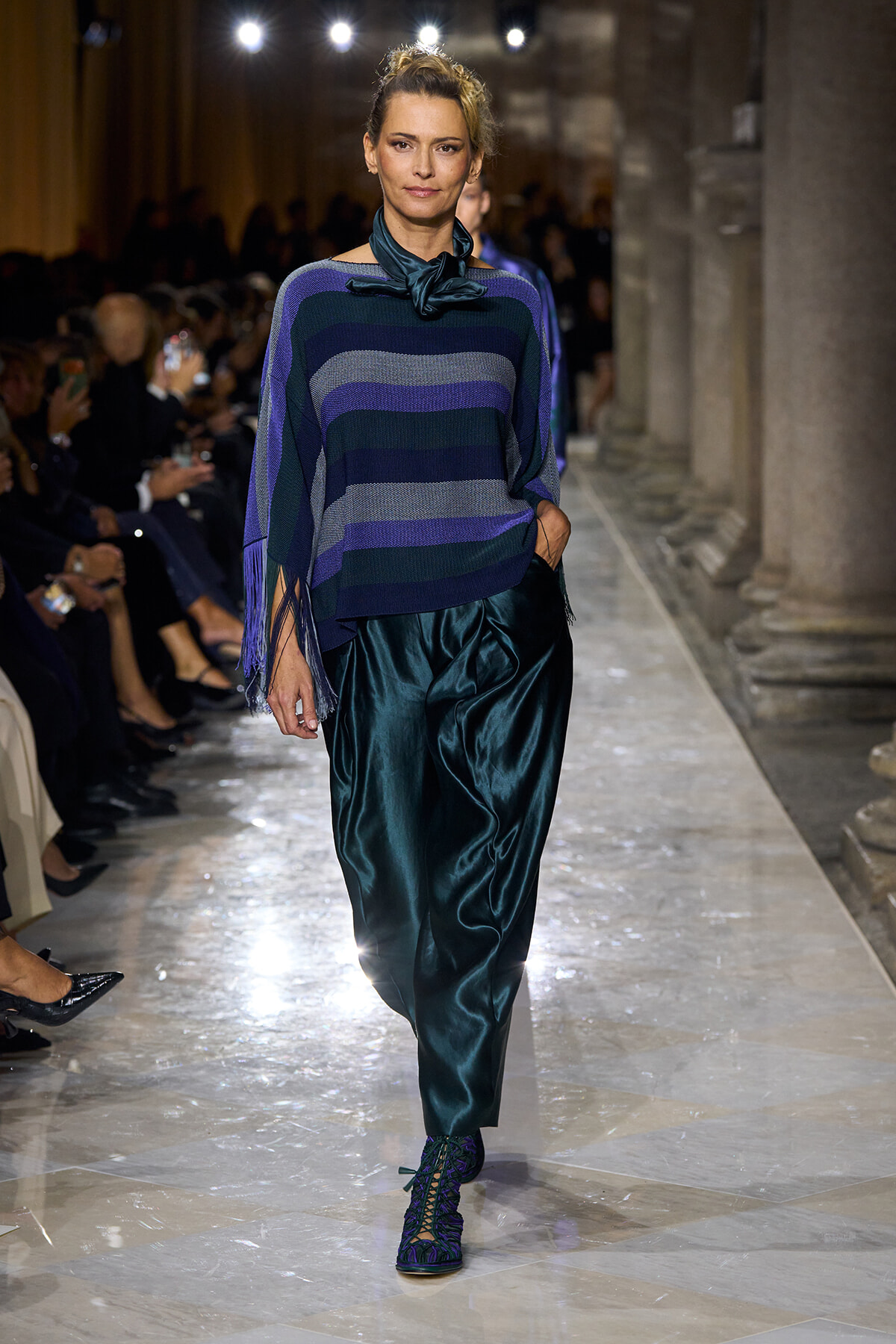 Model on a runway wearing a striped purple and blue poncho with fringe, teal satin pants, and matching lace-up teal shoes