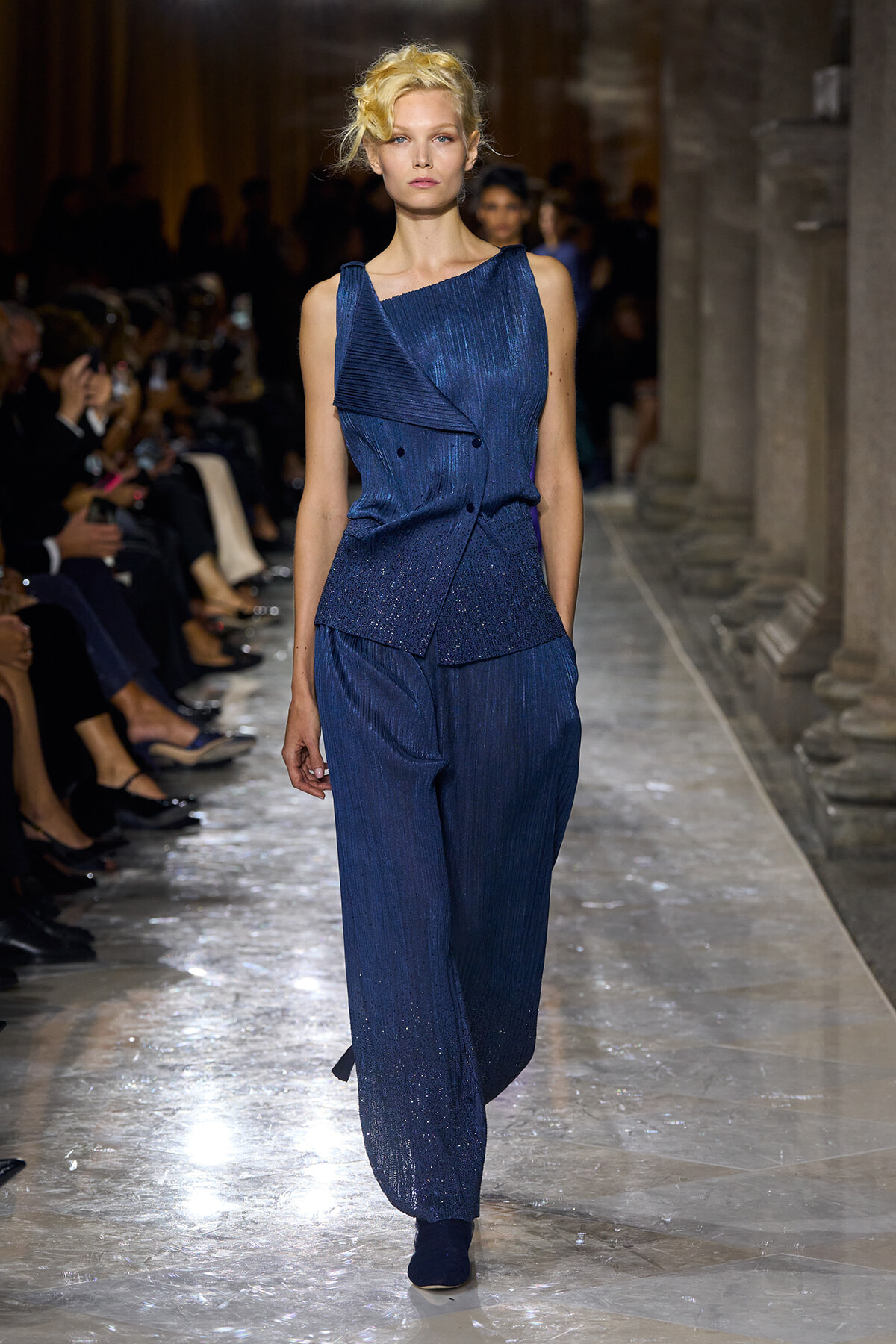 Model walks a runway in a sleeveless, deep blue, textured jumpsuit with an asymmetric top and wide-leg pants; metallic shimmer throughout. She wears dark blue shoes and the audience watches from the sides in a grand venue.