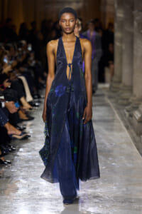 Model walks the runway in a navy halter gown with a deep plunging neckline, tassel ties, and a long flowing, layered skirt on a glossy catwalk.