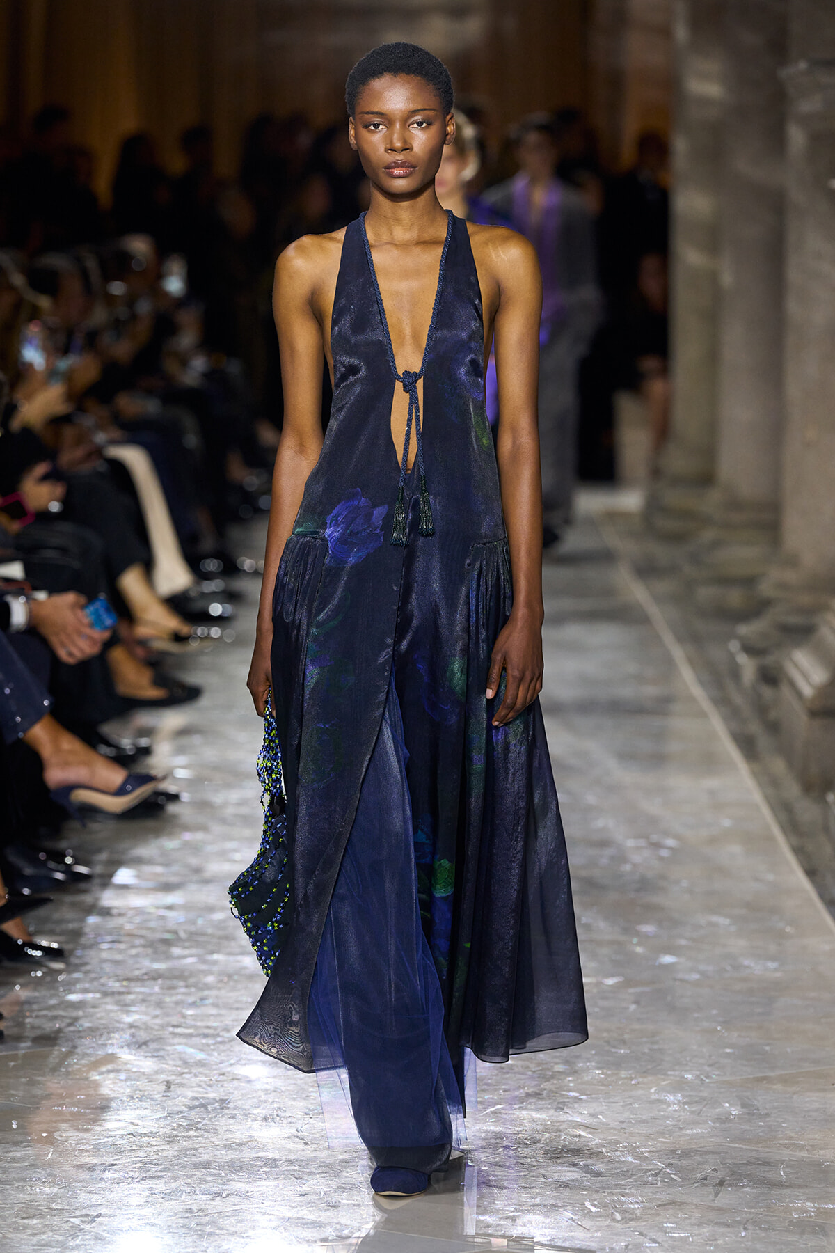Model walks the runway in a navy halter gown with a deep plunging neckline, tassel ties, and a long flowing, layered skirt on a glossy catwalk.