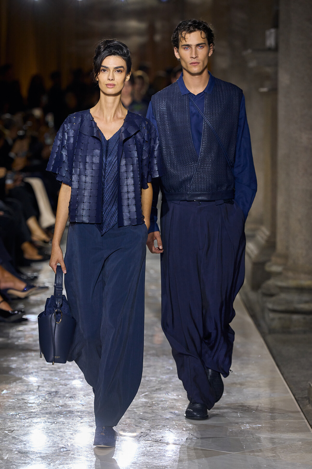 Two models walk a runway in navy blue outfits; the woman on the left carries a matching blue handbag while the man on the right wears a textured vest and wide trousers, both in blue.