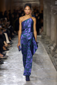 Model walking a runway in a purple-blue tie-dye one-shoulder jumpsuit, holding a small purple clutch.