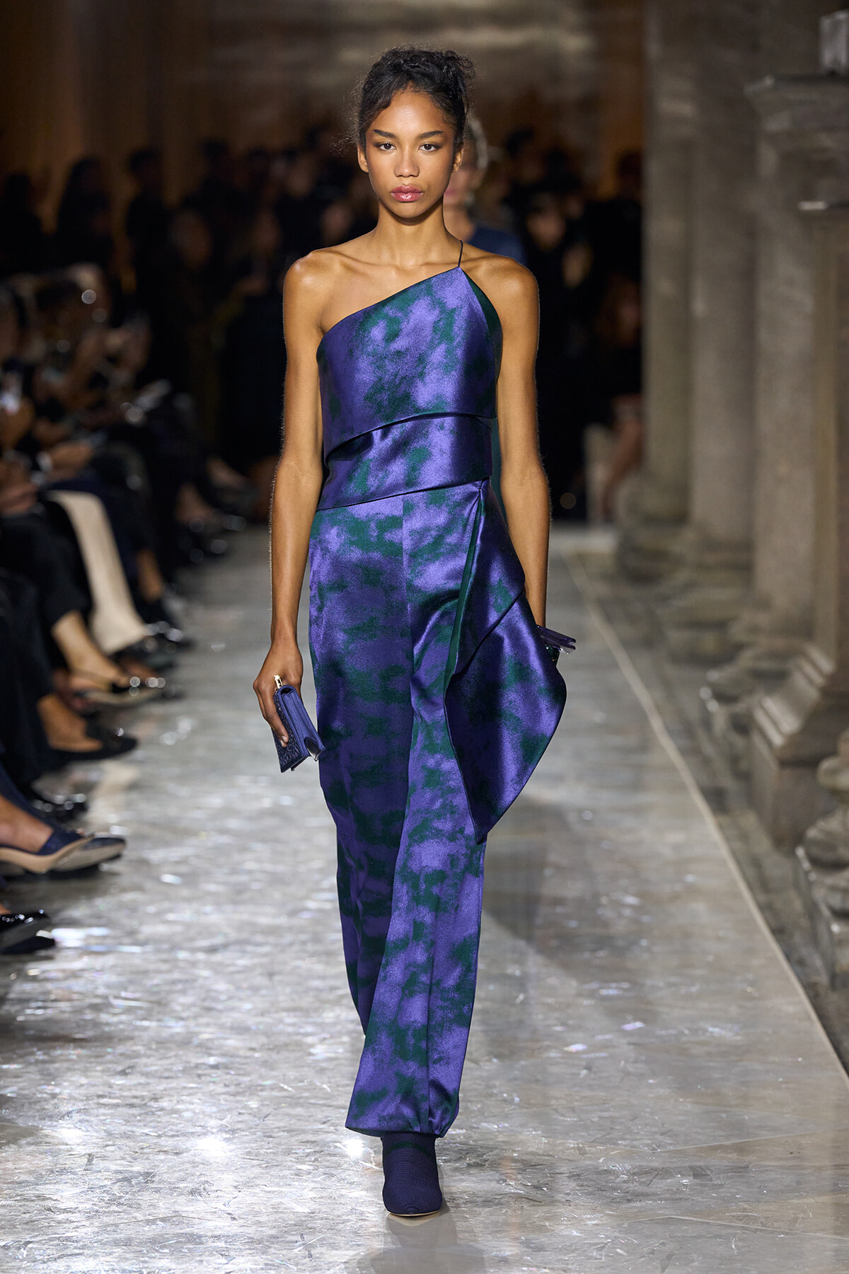 Model walking a runway in a purple-blue tie-dye one-shoulder jumpsuit, holding a small purple clutch.