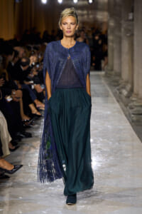 Female model walks a runway in a cobalt blue pleated cape over a dark top and teal wide-leg pants, audience blurred in background