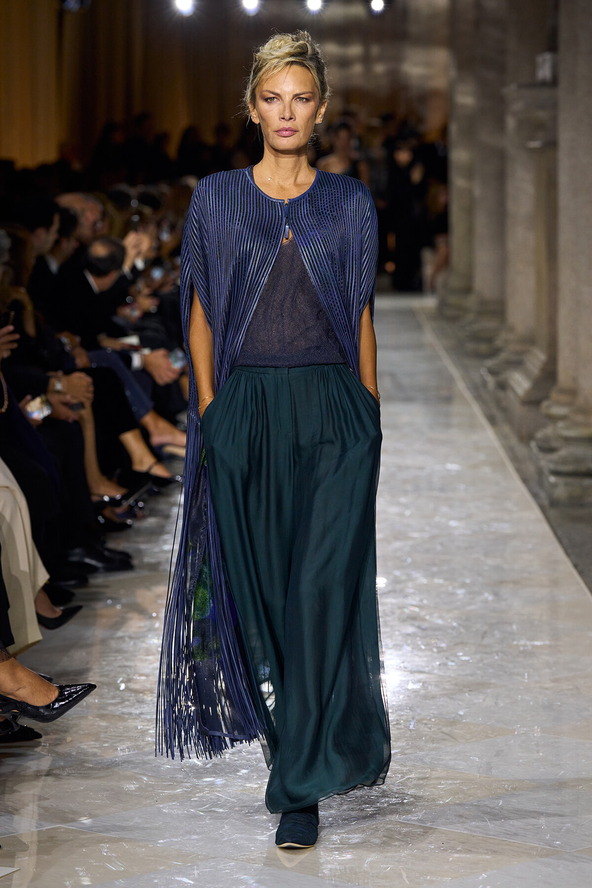 Female model walks a runway in a cobalt blue pleated cape over a dark top and teal wide-leg pants, audience blurred in background