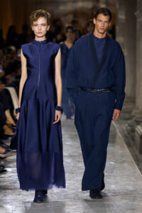 Two models walk a runway in navy-blue outfits: a sleeveless zip-front gown on the left and a loose jumpsuit with a belt on the right.