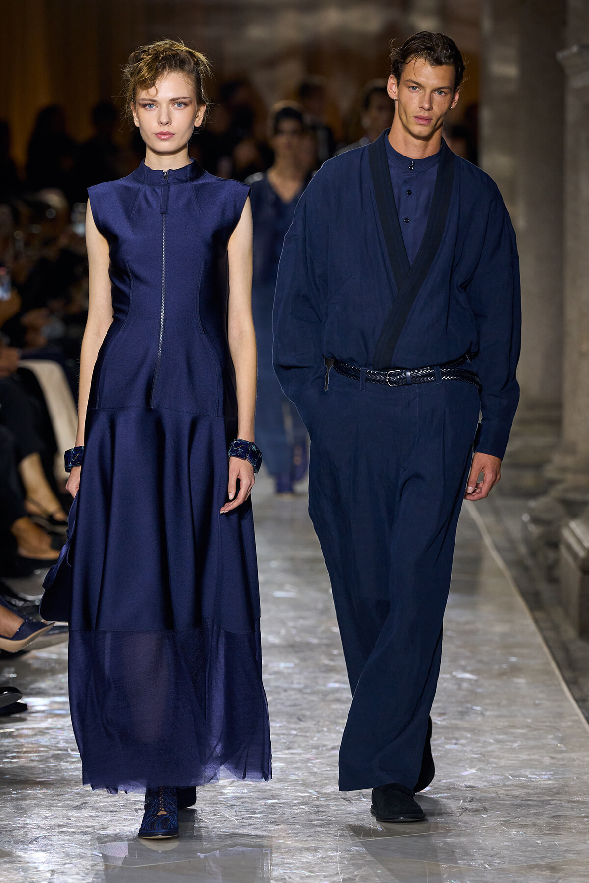 Two models walk a runway in navy-blue outfits: a sleeveless zip-front gown on the left and a loose jumpsuit with a belt on the right.