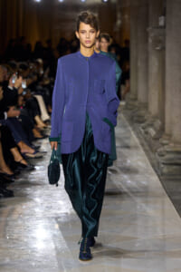Model on a runway wearing a lavender oversized jacket with green cuffs and pockets, teal satin pants, and a small beaded bag.