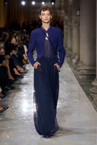 Model walks the runway in a cobalt blue pinstriped jacket over a sheer top and wide navy sheer pants, with a crossbody bag; audience in the background.