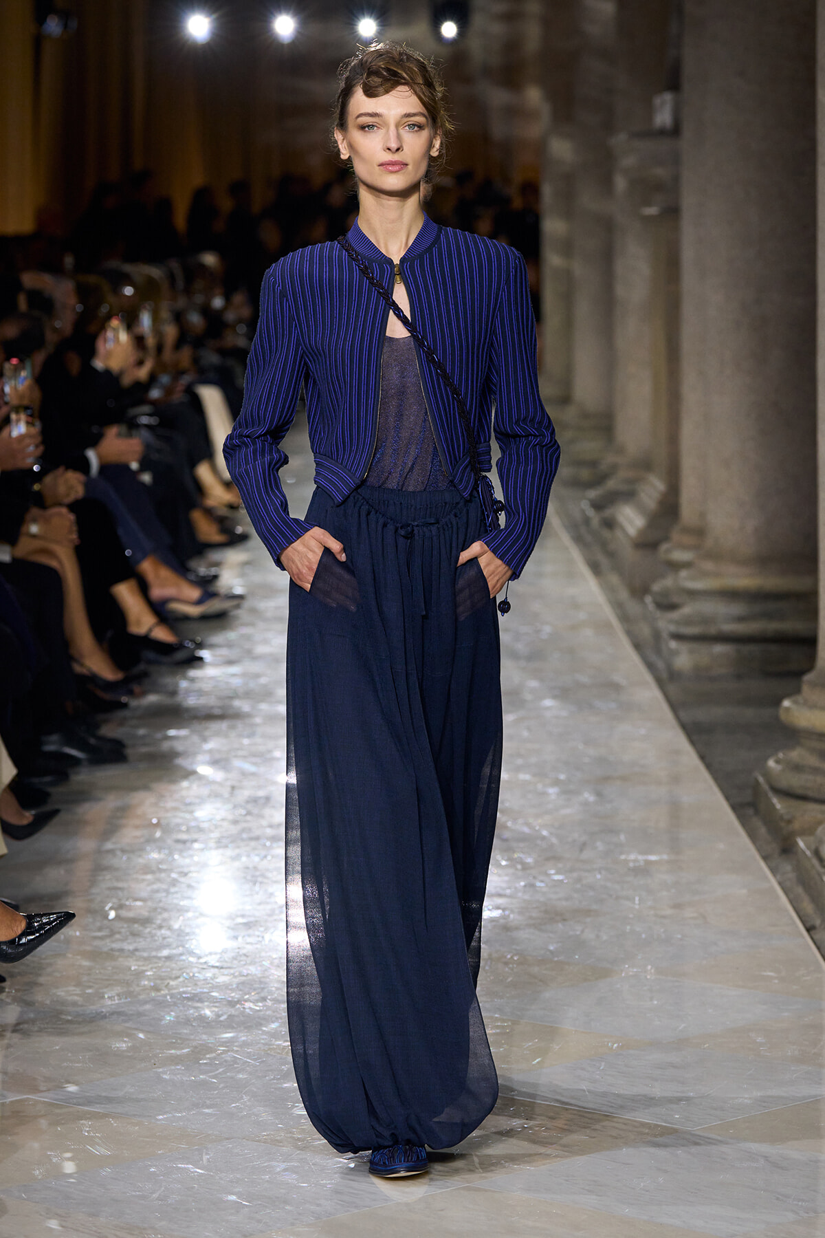 Model walks the runway in a cobalt blue pinstriped jacket over a sheer top and wide navy sheer pants, with a crossbody bag; audience in the background.