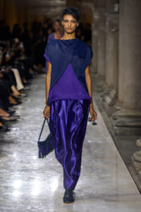 Model walking the runway in a navy and purple color-block outfit with a fringed green handbag on the arm or held at side.