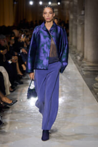 Model walks a runway in a cobalt-purple satin jacket over a sheer top and wide-leg purple trousers, carrying a fringed beaded handbag.
