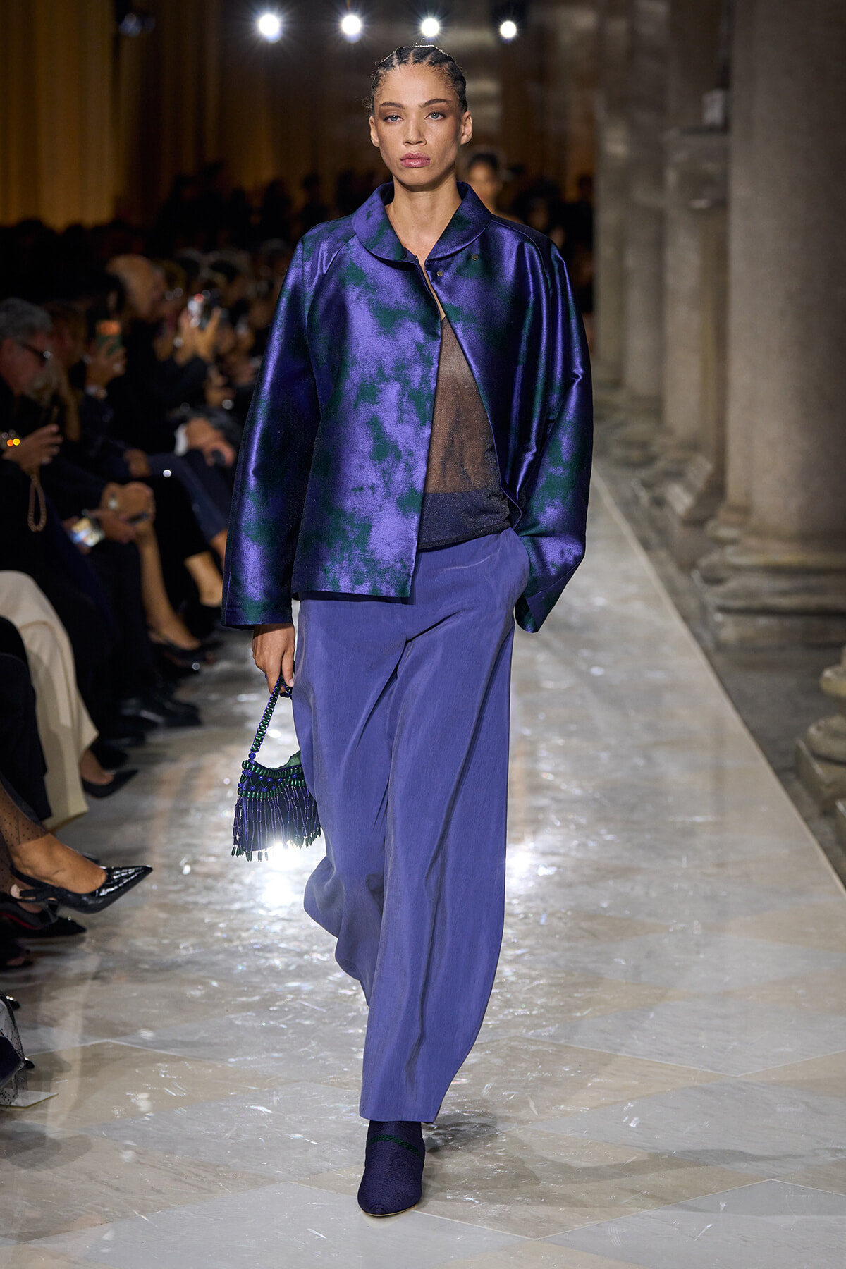 Model walks a runway in a cobalt-purple satin jacket over a sheer top and wide-leg purple trousers, carrying a fringed beaded handbag.
