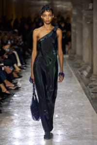 Model walks a fashion runway in a one-shoulder black satin gown with subtle blue floral accents, carrying a blue beaded bag on the left.