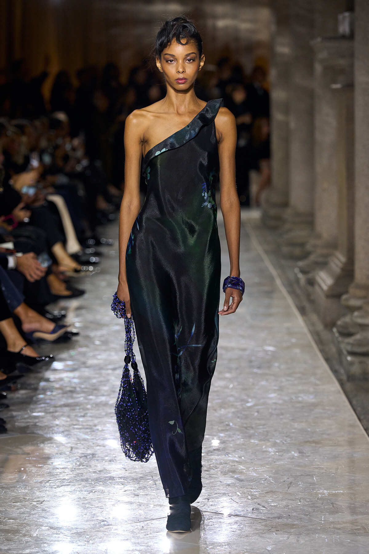 Model walks a fashion runway in a one-shoulder black satin gown with subtle blue floral accents, carrying a blue beaded bag on the left.