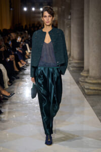 Female model on a fashion runway wearing a teal speckled jacket over a dark sheer top, emerald satin trousers, patterned blue shoes, and a small beaded bag.