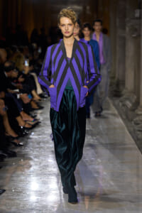 Model walks a runway in a bold purple and black chevron-striped blazer paired with teal velvet pants.