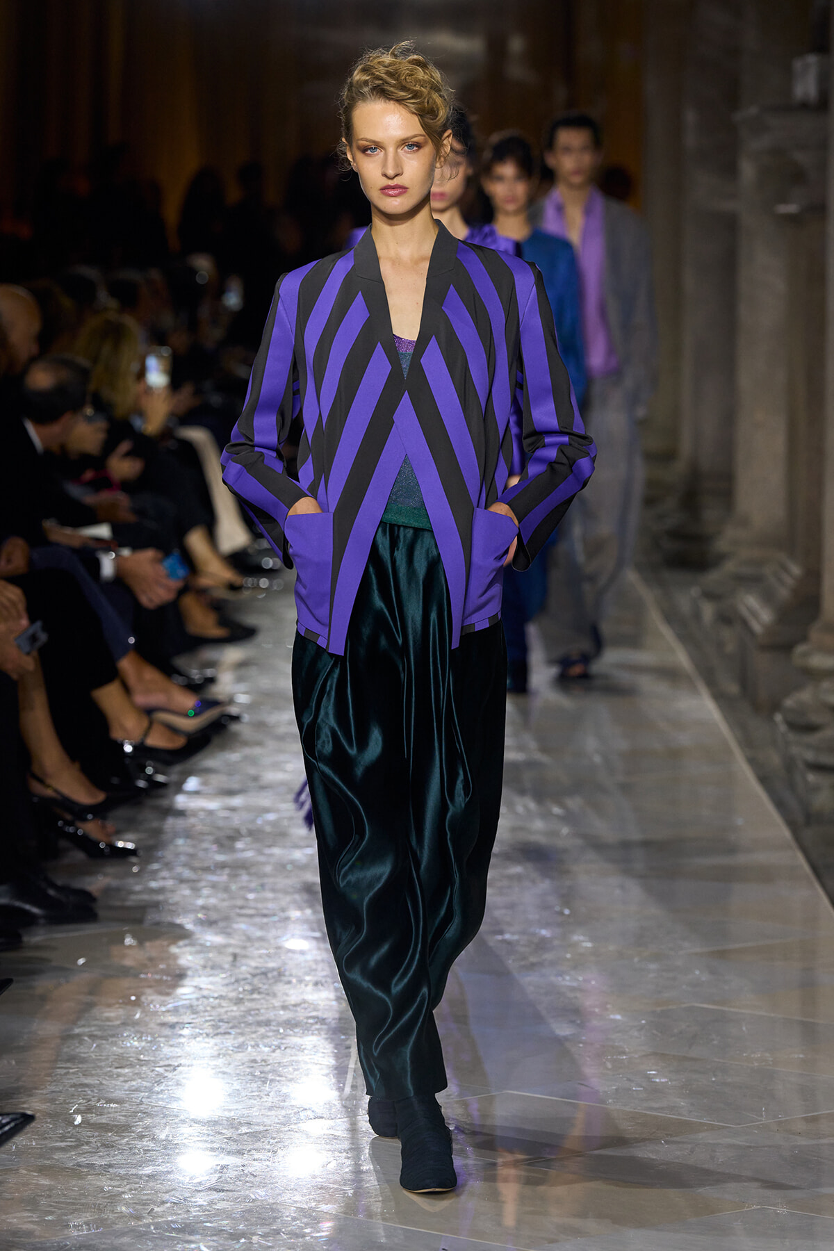 Model walks a runway in a bold purple and black chevron-striped blazer paired with teal velvet pants.