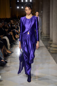 Model on a runway wearing a shiny purple satin wrap blazer with matching pants and a fringed purple handbag.