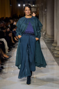 Model walking a runway in teal-blue layered outfit: flowing coat over wide-leg pants with blue top and decorative belt.