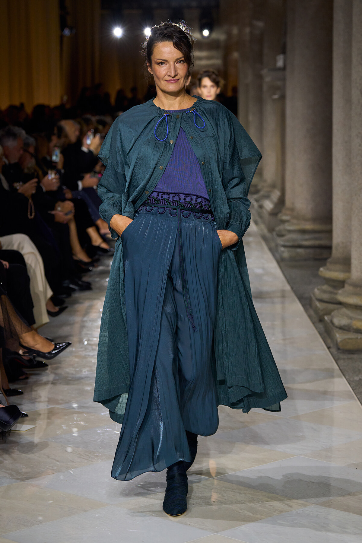 Model walking a runway in teal-blue layered outfit: flowing coat over wide-leg pants with blue top and decorative belt.