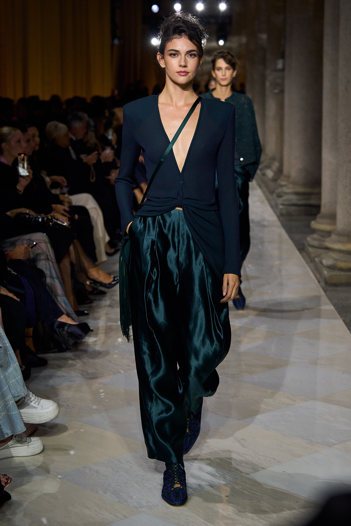 Model walks the runway in a dark teal knit top with a deep plunging neckline and twisted knot at the waist, paired with glossy emerald satin pants; a crossbody bag strap crosses the torso. Audience seated along the catwalk behind.