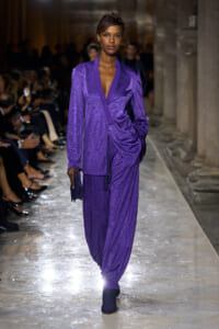 Model walking the runway in a shimmering purple suit with a deep V-neck and draped, ruched pants at a fashion show.