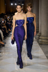 Two female models walk a runway in shimmering blue strapless gowns with waist cutouts, audience watching from sides.
