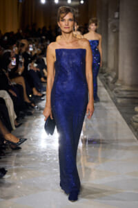 Model walks the runway in a strapless blue sequin gown, holding a matching clutch, with an audience in the background.
