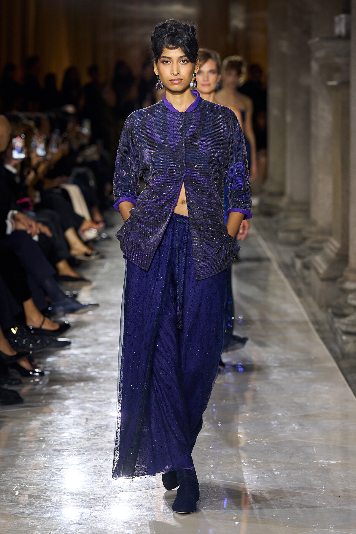 Model walks a runway in a glittery purple ensemble: a sequined jacket over a flowing, sparkly purple skirt with hands in pockets.