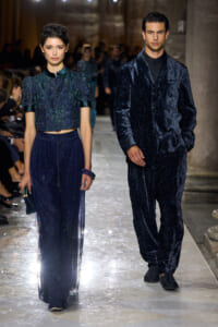 Two models walk a runway in coordinated navy outfits: a woman in a glittery cropped blouse with a sheer floor-length skirt and a man in a matching velvet suit, both in deep blue tones.