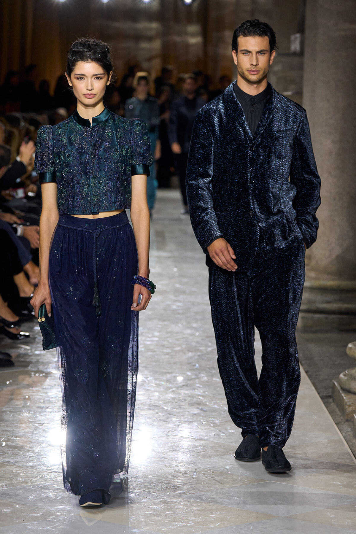 Two models walk a runway in coordinated navy outfits: a woman in a glittery cropped blouse with a sheer floor-length skirt and a man in a matching velvet suit, both in deep blue tones.